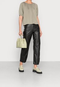 Top khaki a maniche corte con scollatura arricciata, abbinato a pantaloni larghi in pelle nera. La modella tiene in mano una borsa verde chiaro con accessori dorati.