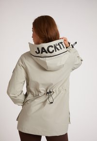 Lichtgrijze waterdichte jas met een hoge kraag, verstelbare trekkoord taille en een groot zwart "JACK1T" logo op de achterkant. Gestructureerde stof.