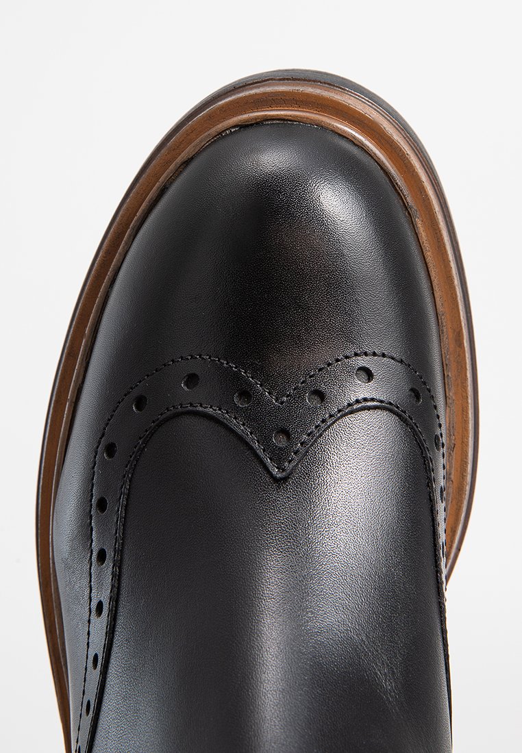 Scarpa di pelle nera con punta arrotondata, caratterizzata da perforazioni decorative e un design curvo. La suola marrone accentua la base.