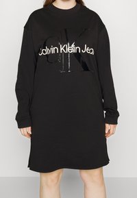 Černé oversize šaty z bavlny se dlouhými rukávy. Obsahují lesklé černé logo "CK" a text "Calvin Klein Jeans" v bílé barvě.