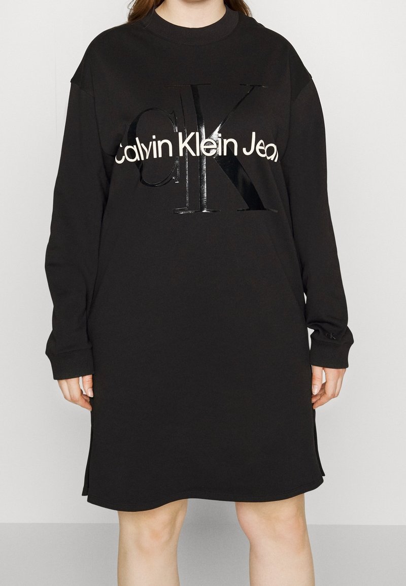 Černé oversize šaty z bavlny se dlouhými rukávy. Obsahují lesklé černé logo "CK" a text "Calvin Klein Jeans" v bílé barvě.