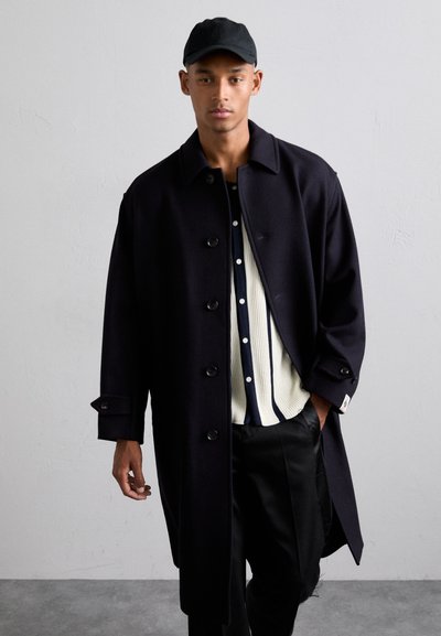 Dlouhý černý vlněný kabát s velkými knoflíky, bočními kapsami a volným střihem, nošený přes bílou pruhovanou košili a černé kalhoty.