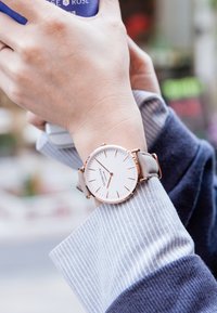 Orologio rotondo con quadrante bianco e dettagli in oro rosa. Presenta un cinturino in pelle grigia e indicatori delle ore sottili. Indossato su una mano con maniche a righe.