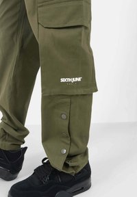 Olijfgroene cargobroek met een zak en zilveren drukknoopjes bij de zoom, gecombineerd met zwarte sneakers met een gestructureerd ontwerp.