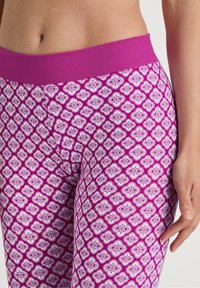 Close-up van een persoon die paarse leggings draagt met een wit geometrisch bloempatroon en een effen paarse tailleband.
