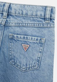 Jeans de mezclilla azul claro con un bolsillo trasero triangular que presenta un parche con el logo en rojo y blanco de "GUESS", tejido texturizado y detalles de costura clásica.