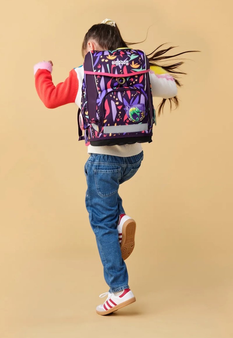Child jumping with colorful floral-patterned backpack, wearing red and white sweater, blue jeans, and white sneakers with red stripes.