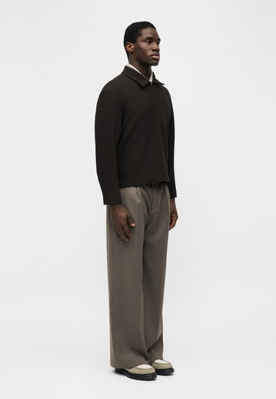 Άνδρας που στέκεται φορώντας μαύρο μπουφάν με φερμουάρ, παντελόνι φαρδύ σε ταουπέ χρώμα, και μπεζ-μαύρα παπούτσια μπροστά σε απλό λευκό φόντο.