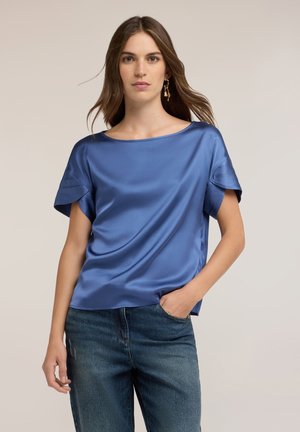 Vrouw met lang bruin haar, gekleed in een blauw satijnen top met korte mouwen en blauwe jeans, staand met één hand in de zak tegen een beige achtergrond.
