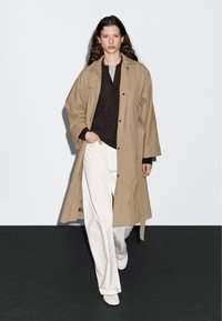 Beige lange jas met een ceintuur in de taille, over een donkere trui. Gepaard met witte wijde broek en lichte schoenen. Glad stof, relaxte pasvorm.