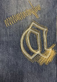 Denim azul con intrincados bordados en amarillo y blanco que forman una letra estilizada y patrones en zigzag, añadiendo textura y profundidad.