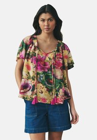 Bloem blouse in geel, met roze en groene patronen, korte mouwen, een V-hals en franjes. Gecombineerd met blauwe denim shorts.