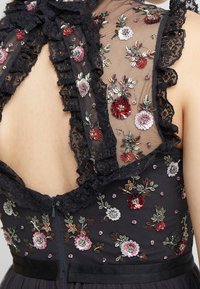 Robe noire avec un dos transparent brodé de motifs floraux, des sequins et des volants en dentelle le long de l'encolure. Détail de fermeture éclair.