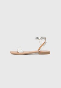 Zilveren platte sandalen met een metallic band, een tan zool en een enkelbandje met kettingdetail. Eenvoudig ontwerp met minimale accenten.