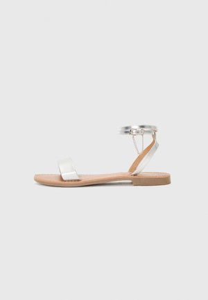 Sandales plates argentées avec une bride métallique, une semelle beige et une bride de cheville ornée d'un détail en chaîne. Design simple avec des accents minimalistes.