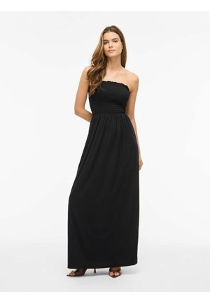 Vrouw met lang bruin haar, die een strapless zwarte maxi-jurk en bruine open-toe hakken draagt, staand voor een effen witte achtergrond.