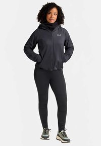 Zwarte gewatteerde jas met capuchon en een volledige ritssluiting, met een gladde textuur en logodetail. Draagt over zwarte leggings en wandelschoenen met gestructureerde zolen.