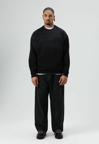 Zwart oversized sweatshirt met een gestructureerd oppervlak, gedragen over een wit T-shirt, gecombineerd met losse zwarte jeans en zwarte schoenen met roze veters.