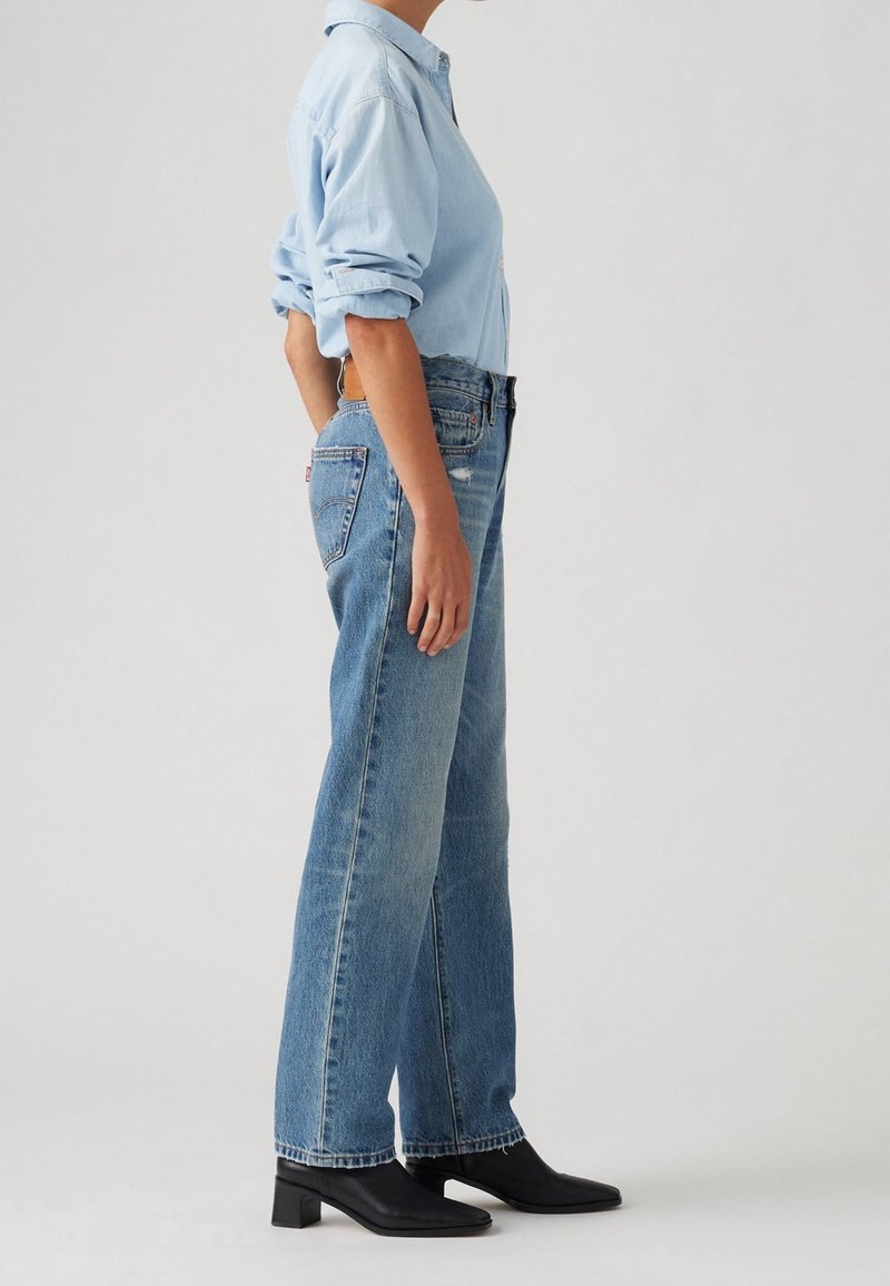 Personne en chemise bleu clair retroussée, jean bleu taille haute et jambe droite, et bottines noires à talons, debout de profil.