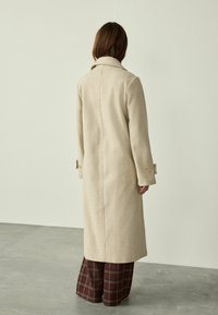 Abrigo de lana color beige con cuello estructurado, mangas largas y detalles de botones. Llevado sobre pantalones de cuadros de pierna ancha. Diseño simplistic.