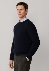 Homme portant un pull côtelé bleu marine par-dessus une chemise habillée bleu clair, debout avec une main dans la poche contre un fond clair uni.