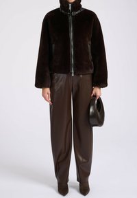 Bruine nepbontjas met een hoge kraag, rits aan de voorkant en leren accenten. Gecombineerd met bruine leren broeken en een ronde handtas.