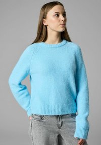 Jeune femme portant un pull bleu vif en matière pelucheuse et un jean gris clair, regardant légèrement vers le haut et vers la droite, la main dans la poche.