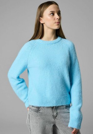 Young woman wearing a bright blue fuzzy sweater and light gray jeans, looking slightly upward and to the right with her hand in her pocket.