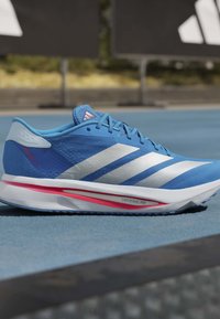 Μπλε παπούτσι για τρέξιμο Adidas με λευκές ρίγες και κόκκινες λεπτομέρειες, ακουμπισμένο σε μπλε επιφάνεια στίβου.