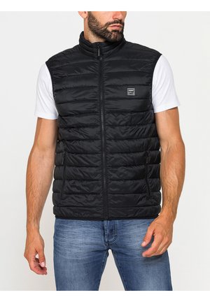 Uomo che indossa un gilet nero imbottito senza maniche sopra una T-shirt bianca e jeans blu, in piedi davanti a uno sfondo chiaro uniforme.