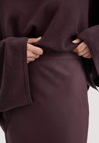 Handen die een donkerpaarse oversized trui met wijde mouwen aanpassen over een bijpassende hoge taille rok op een lichte achtergrond.