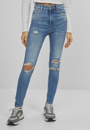 Femme portant un jean skinny bleu clair déchiré, un haut blanc crop à manches longues, et des baskets grises, debout devant un fond uni.
