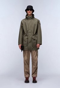 Olivgrüne wasserdichte Parka mit tailliertem Schnitt, zwei Fronttaschen, kombiniert mit beigen Cargohosen und braunen Schnürstiefeln, Accessoires umfassen Sonnenbrille und Fischerhut.