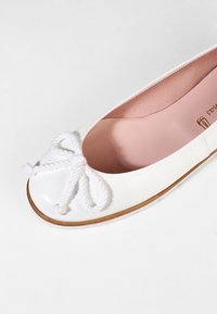 Chaussure ballerine plate blanche avec un bout arrondi, un nœud tressé blanc sur le dessus, une semelle beige et une doublure intérieure rose.