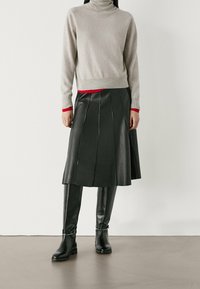 Grauer Rollkragenpullover, tailliert, kombiniert mit einem schwarzen Leder-Faltenrock. Kniehohe schwarze Stiefel mit silbernen Schnallenakzenten.