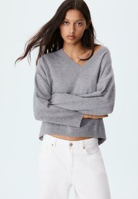 En ung kvinde med langt mørkt hår iført en grå V-hals sweater og hvide bukser, med armene foldet, mod en ensfarvet lys baggrund.