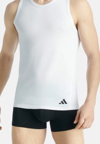 Bílý bezramenný top vyrobený z hladké látky, s černým logem Adidas v levém dolním rohu. Nosený s černými úzkými šortkami.