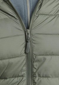 Olivgrön pufferjacka med horisontellt quiltat mönster, försett med en metalldragkedja och en ljusgrå innerlining. Mjuk struktur och lätt design.