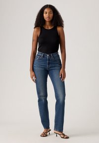 Svart ribbestrikket tanktopp sammen med høytlivede, litt falmede blå denimjeans. Mørke hælsandaler fullfører antrekket.