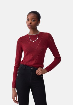 Giovane donna che indossa un maglione rosso strutturato con motivo a V, pantaloni neri a vita alta con bottoni e collane d’argento a strati.