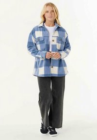 Camicia di flanella a quadri blu e bianca con chiusura a bottoni, due tasche sul petto e vestibilità leggermente oversize. Abbinata a pantaloni neri a gamba larga.