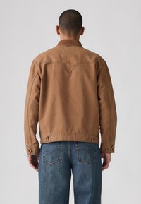 Bruine katoenen jas met een rugverliesontwerp, drukknoopsluitingen bij de manchetten en gecombineerd met blauwe denim jeans. Heeft een soepele textuur.
