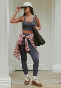 Top corto sportivo grigio e leggings a vita alta abbinati a una camicia a righe rossa e bianca annodata in vita, con una borsa marrone.