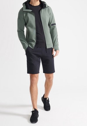Mann trägt grünen Kapuzenpullover mit Reißverschluss, schwarzes T-Shirt, schwarze Shorts und schwarze Sneakers, steht mit den Händen in den Taschen vor weißem Hintergrund.