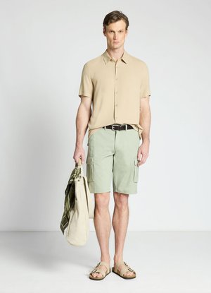 Uomo con camicia beige a maniche corte, pantaloncini cargo verde chiaro, sandali, che tiene in mano una borsa beige con sciarpa verde su sfondo bianco.