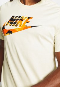 Ljusbeige bomulls-T-shirt med en grafisk Nike-logotyp i orange, svart och gula nyanser med en texturerad, mönstrad design.