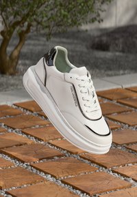 Λευκό δερμάτινο αθλητικό παπούτσι με μαύρες λεπτομέρειες, στρογγυλεμένη μύτη, λευκά κορδόνια και διακριτικό λογότυπο. Διαθέτει μαλακή σόλα και λεία υφή.