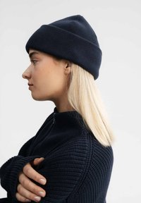 Gebreide marineblauwe beanie met een ribbelstructuur, voorzien van een opgevouwen rand. Draag door een persoon met steil blond haar en een marineblauwe hoge halstrui.