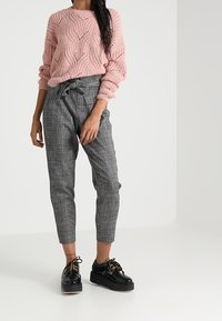 Kobieta ubrana w różowy, teksturowany sweter, szare spodnie w kratę z wysokim stanem i paskiem w talii oraz czarne, sznurowane buty na platformie.