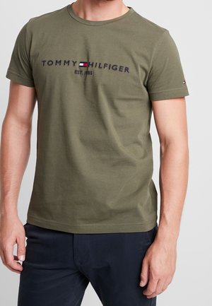 Muž v olivově zeleném tričku Tommy Hilfiger s logem a námořnických kalhotách na jednobarevném pozadí.